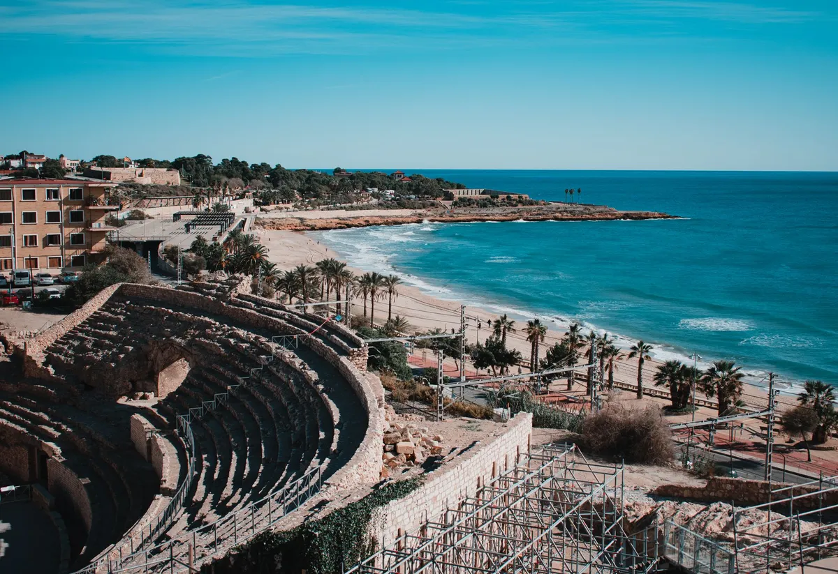 El anfiteatro romano de Tarragona sobre la playa del Miracle, con las palmeras del paseo, la vía del tren pegada a la arena y el Mediterráneo al fondo.
